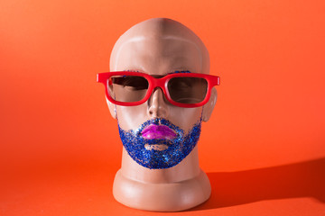 Mannequin male head in red glasses with blue glitter beard and eyebrowns and pink lipstick. Male...