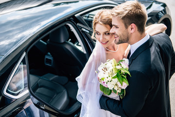 handsome bridegroom in suit hugging attractive and blonde bride with bouquet
