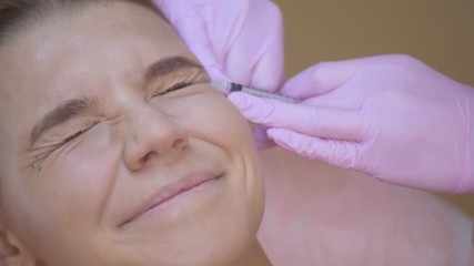 The doctor beautician makes injections in the face of the female patient. concept of beauty and cosmetology. 4k. biorevitalization The patient squints and smiles.