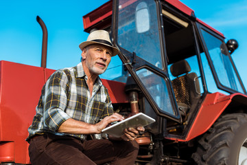 Fototapeta na wymiar senior farmer in straw hat using digital tablet near red tractor