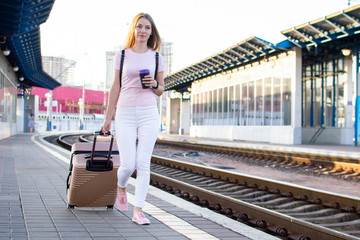 attractive girl is standing with luggage at the station and waiting for the train, the student is going on a trip, she is walking along the platform, copy space