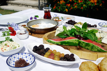 A breakfast table composed of cheese, chocolade cream, milk cream, olives and various marmalades, placed in front of flowers at a garden