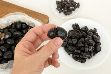 Woman hand holds black olive crushed with a knife for pitting it on a background of kithen table with plate and chopping board. Homemade black olive paste with anchovies, garlic and olive oil.