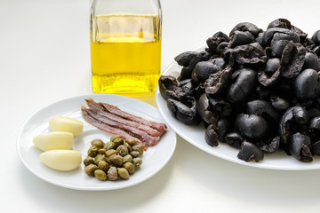 Ingredients for tapenade: pitted black olives on white plate, olive oil in glass bottle, three cloves of garlic, some anchovy fillets and pickled capers on a sauser. Healthy eating.