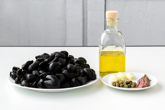 Tapenade ingredients: pitted black olives on white plate, olive oil in glass bottle, three cloves of garlic, some anchovy fillets and pickled capers on a sauser. Healthy eating.