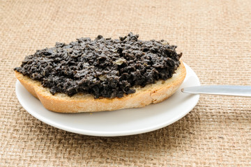 Fresh tapenade sandwich with slice of wheat bread in a white sauser and table knife on a burlap table napkin. Homemade black olive paste with anchovies, garlic and olive oil.