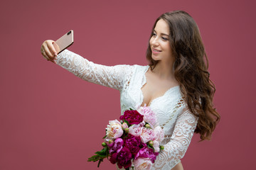 Beautiful pregnant woman in lace white lingerie body suit makes a selfie with a bouquet of flowers peony