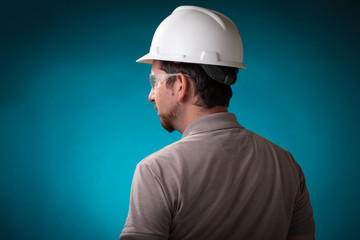 Construction worker with beard and white safety helmet seen from the back with protective glasses on against a studio background and blue spotlight behind