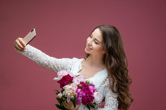 Beautiful pregnant woman in lace white lingerie body suit makes a selfie with a bouquet of flowers peony