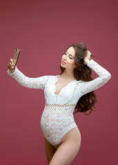 Beautiful pregnant woman with long hair dressed in lace white lingerie with a body suit doing a selfie on a pink background in the studio