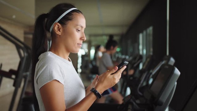 Attractive Young Mixed Race Girl Using Fitness App on Mobile Phone in the Gym. Health Sport Motivation Concept. 4K Slowmotion.