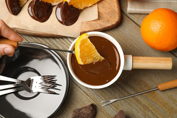 Woman dipping slice of orange into fondue pot with milk chocolate at wooden table, top view