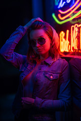 Sexy portrait of a young girl with an open bust in sunglasses and with in the night city, with creative light on the background of neon lamps.Night clubs, parties, strip business, night life.Vertical