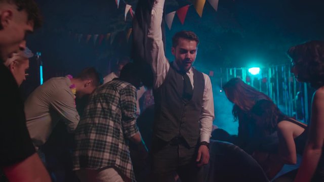 Handsome sexy businessman in suit walking like boss throwing money on young people dancing with modern moves into electronic music at club party.