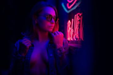 Fototapeta na wymiar Sexy portrait of a young girl with an open bust in sunglasses and with in the night city, red-blue light of neon lamps. Night clubs, parties, strip business, night life, business game. Banner