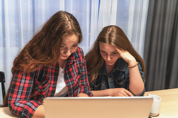 Two attractive young women at home with laptop, talking with each other and laughing. Friendship or female couple concept