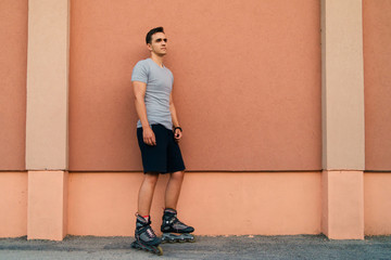 Portrait of young man in roller blade skates standing in front of the plain wall leaning wearing gray t shirt and shorts in summer day