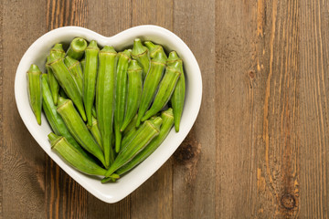 Fresh organic green okra in a heart shaped bowl isolated on a brown wood board with copy space.