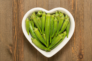 Fresh organic green okra in a heart shaped bowl isolated on a brown wood board.