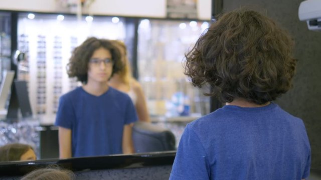 choice of frames for glasses in optics. Mom and her teenager son choose a frame for glasses while looking in a large mirror in optics.