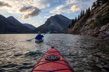 Adventurous Man Kayaking in Glacier Lake surrounded by the beautiful Canadian Rocky Mountains during a cloudy summer sunset. Taken in Upper Waterton Lake, Alberta, Canada.