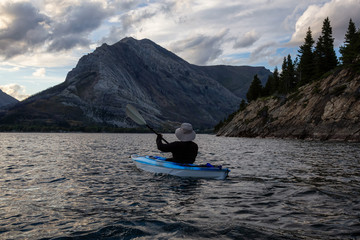 Adventurous Man Kayaking in Glacier Lake surrounded by the beautiful Canadian Rocky Mountains during a cloudy summer sunset. Taken in Upper Waterton Lake, Alberta, Canada.