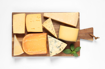 Wooden board with different kinds of cheese and basil on white background, top view