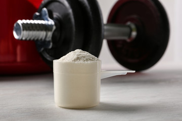 Measuring scoop of protein powder and dumbbell on white table, closeup