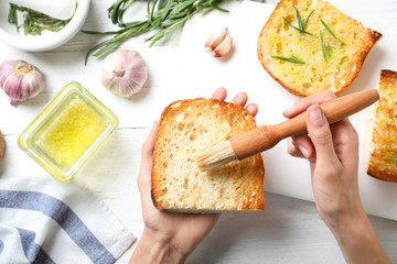 Woman brushing slice of toasted bread with garlic and herb over white wooden table, top view