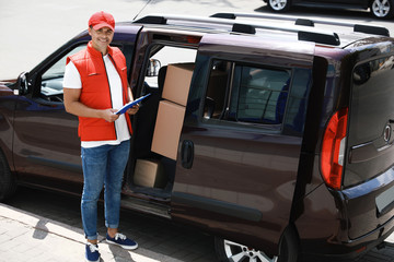 Young courier with clipboard near delivery car outdoors