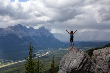 Adventurous Caucasian Girl with open arms is on top of rocky mountain during a cloudy and rainy day. Taken from Mt Lady MacDonald, Canmore, Alberta, Canada.