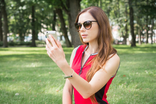 beautiful female in casual wear taking pictures of herself on mobile phone front camera while resting outdoors in park