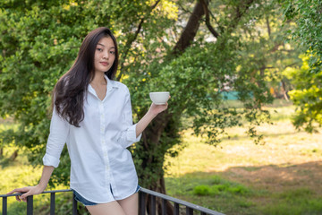 Selective focus of young pretty asian woman standing on balcony in the morning and drinking coffee from coffee cup for breakfast. Cute teenage girl relax on home balcony to get fresh air and sunlight