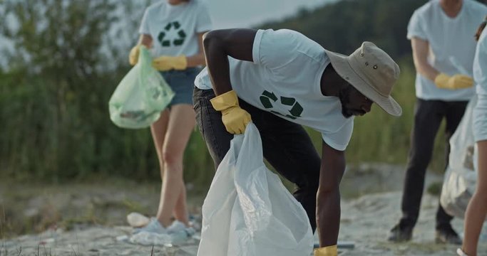 Handsome afro-american guy in eco T-shirt collecting garbage on sandy beach in plastic bags with a group of friends. Volunteerism concept. Anti-pollution operation.