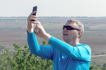 A man in blue longsleeve takes a selfie against the backdrop of a beautiful landscape. Bad cellular signal outside the city, a white man trying to catch a cellular signal