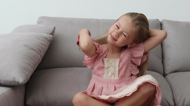 cheerful girl of six years old in a pink dress sits on a sofa and brushes her blonde hair, a girl in a good mood