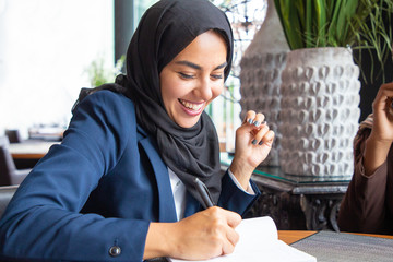 Happy businesswoman taking notes in coffee shop. Young Muslim business woman in hijab and office suit writing in notebook and laughing. Muslim business professional concept