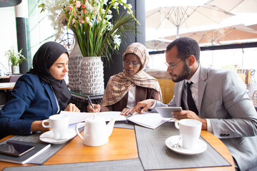 Multicultural business partners discussing contract in cafe. Businessman and Muslim businesswomen...
