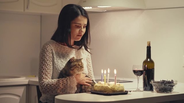 Sad woman sits in the kitchen with a cat and blows out the candles on the cake. Lonely woman celebrates a birthday.