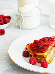 Pancakes with jam are located on a white plate, around the pancakes on a plate are fresh berries. Next to a glass of milk and sour cream. Breakfast, festive food, christmas. Gray background. Close-up.