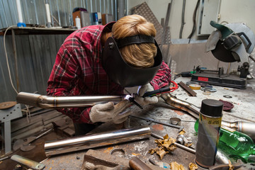 Young guy welder in a checkered red shirt welds a stainless steel pipe for car exhaust using agronomic welding to protect his eyes with a mask in an iron workshop. Modern welding methods.
