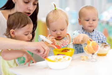 Group of nursery babies eating healthy food with kindergartener help. Lunch break in creche. Little...