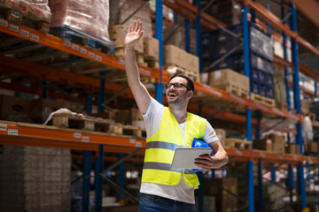 Warehouse worker with tablet and protective uniform standing between shelves in storage center and waving to his coworker.