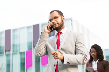 Serious Latin businessman speaking on cell while walking down city street. Confident man in office suit talking on cellphone, city building and passerby in background. Digital technology concept