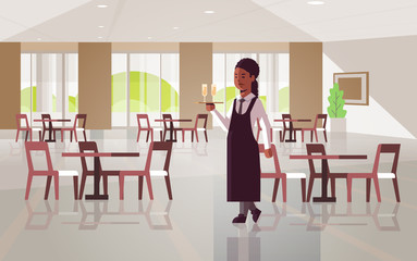 professional waitress holding serving tray with two glasses of champagne african american woman restaurant worker in apron carrying alcohol drinks modern cafe interior flat full length horizontal