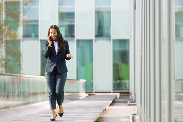 Happy cheerful Latin businesswoman talking on phone while walking outdoors. Young woman in office suit going down urban walkway and calling on cell. Business phone talk concept