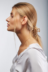 Close-up portrait shot of a young blonde European lady with a goldish hair claw in her long straight hair. The curved hair clip is made of the thick wire. The girl is dressed in a white shirt  