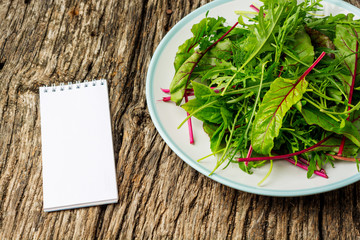 Fresh salad plate with mixed greens (arugula, mesclun, mache) on dark wooden background with notepad close up. Healthy food. Green meal. Flat lay