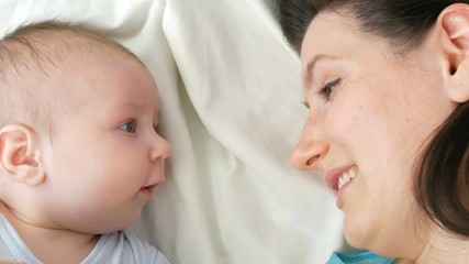 Young beautiful mother with long dark hair is lying on a white bed and playing with a newborn baby of two months