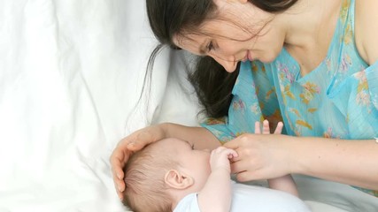 Young beautiful mother with long dark hair is lying on a white bed and playing with a newborn baby of two months